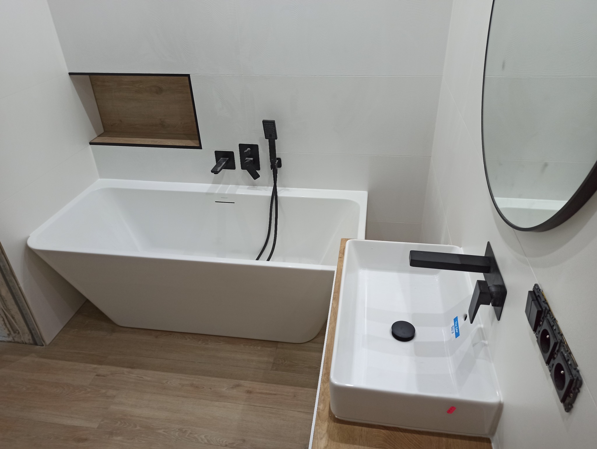 Nowoczesna łazienka z białą wanną, umywalką i czarną armaturą. Wnęka z drewnianym akcentem i okrągłe lustro dodają elegancji. Minimalistyczny design i jasne oświetlenie.