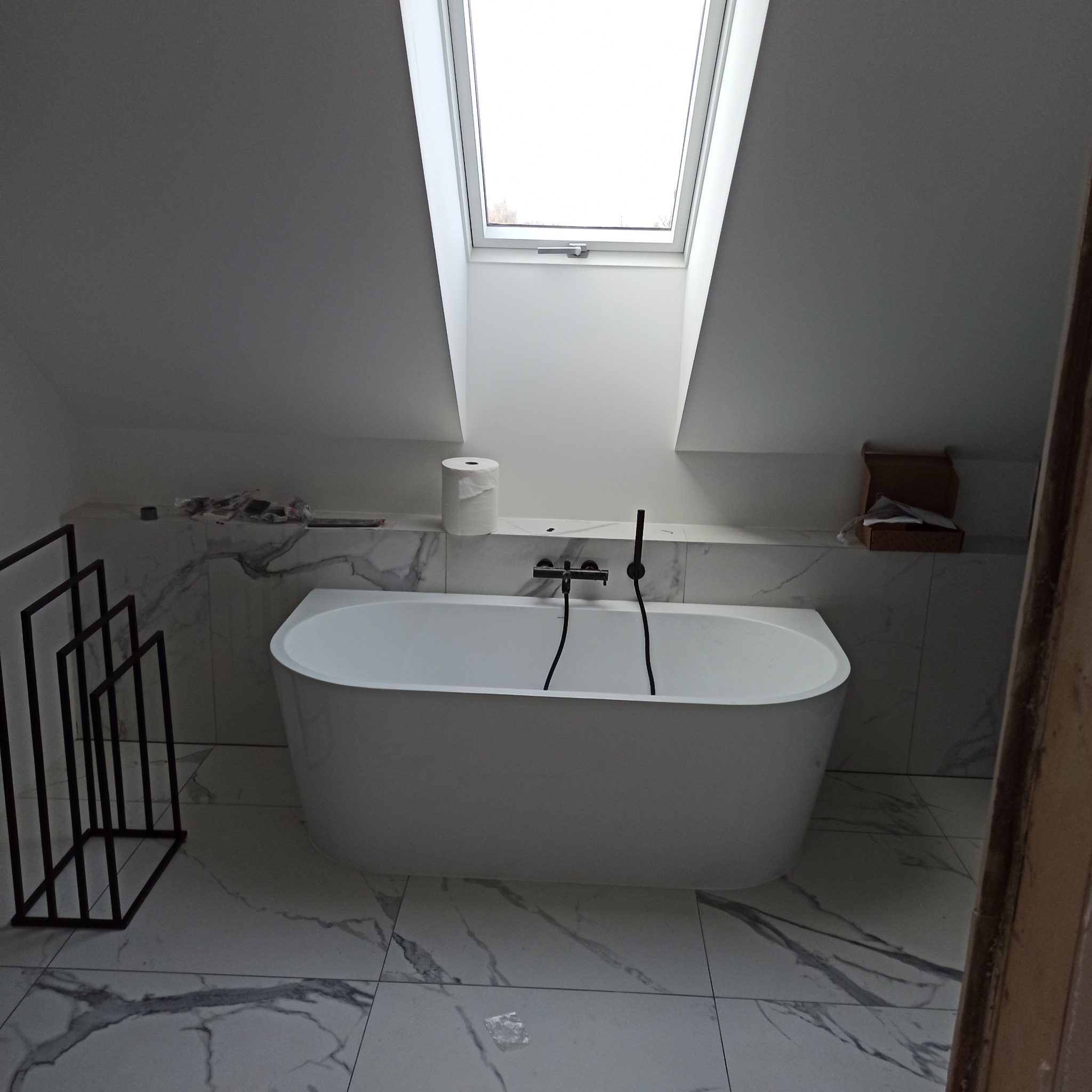 Biała wanna wolnostojąca z czarną armaturą w łazience na poddaszu z oknem dachowym. Marmurowe płytki podłogowe i ścienne. Minimalistyczny wieszak na ręczniki.