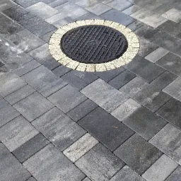 Szara kostka brukowa ułożona w regularny wzór, z okrągłą studzienką kanalizacyjną otoczoną jasnymi kostkami, widoczny fragment ogrodzenia i podlewanie kostki.