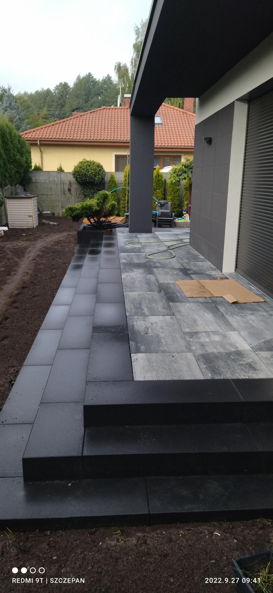 Nowoczesny taras z kostki brukowej w dwóch odcieniach szarości, z niskimi schodami i rabatą z bonsai, obok fragmentu domu z szarą elewacją i roletami.