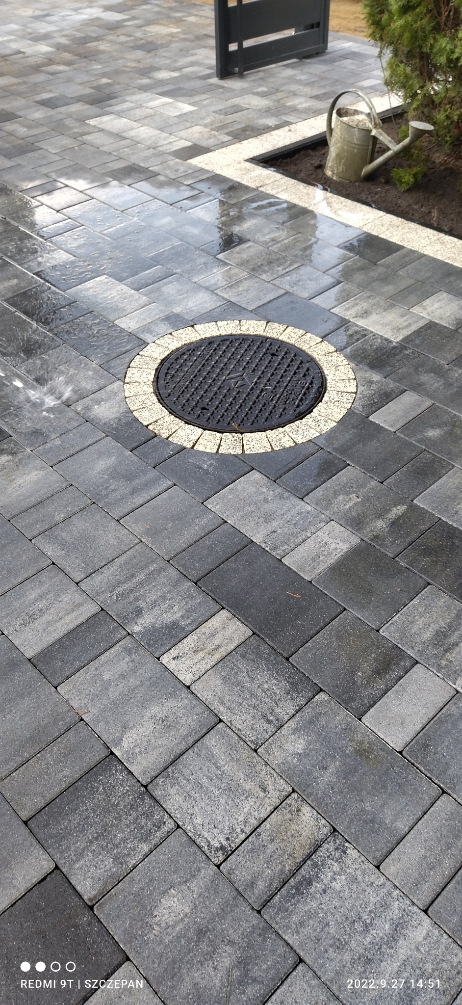 Szara kostka brukowa ułożona w regularny wzór, z okrągłą studzienką kanalizacyjną otoczoną jasnymi kostkami, widoczny fragment ogrodzenia i podlewanie kostki.