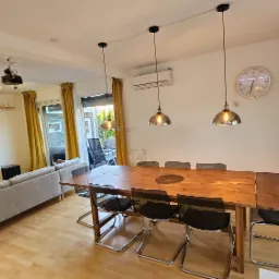 Jasne wnętrze salonu z drewnianą podłogą, długim stołem jadalnym z czarnymi krzesłami, szarą sofą i telewizorem, oświetlone trzema wiszącymi lampami i światłem wpadającym przez okno balkonowe...