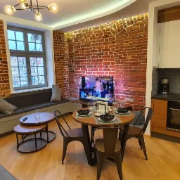Wnętrze mieszkania z ceglaną ścianą, drewnianą podłogą i aneksem kuchennym; stół nakryty do posiłku, telewizor włączony, widok z okna na zabytkową architekturę.