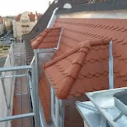 Widok z góry na fragment dachu z czerwonej dachówki karpiówki i metalowe obróbki blacharskie, widoczne okna dachowe, rynna i fragment rusztowania, w tle zabudowa miejska.