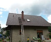 Dom jednorodzinny z nowym, brązowym dachem z dachówki ceramicznej, widoczny komin i okno dachowe, drabina oparta o dach, elewacja budynku częściowo otynkowana, w tle zieleń i błękitne niebo...