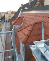 Widok z góry na fragment dachu z czerwonej dachówki karpiówki i metalowe obróbki blacharskie, widoczne okna dachowe, rynna i fragment rusztowania, w tle zabudowa miejska.