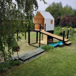 Drewniany plac zabaw z domkiem na podeście, mostkiem linowym i małą zjeżdżalnią, otoczony zielonym trawnikiem i drzewami, widok z góry.