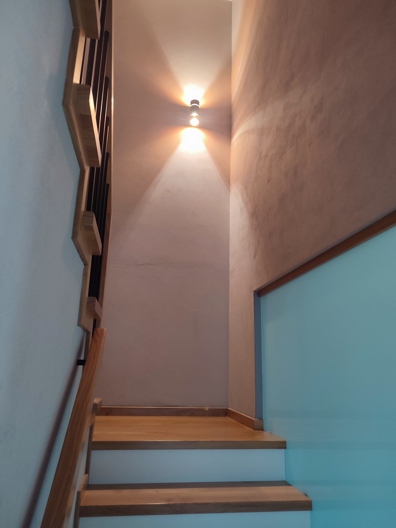 Klatka schodowa z drewnianymi schodami i balustradą, ściana z efektem dekoracyjnym w odcieniach beżu i oświetleniem punktowym.