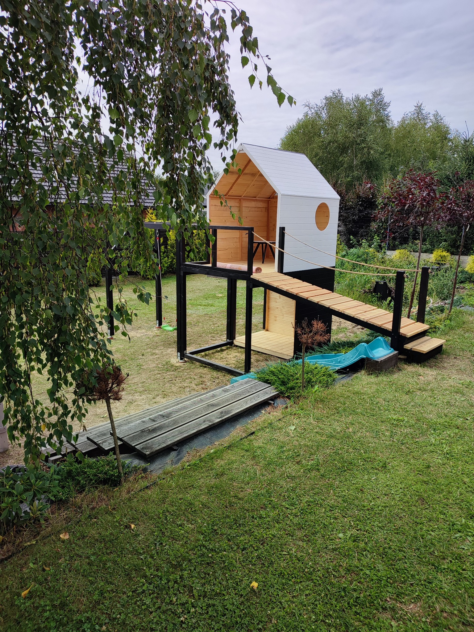 Drewniany plac zabaw z domkiem na podeście, mostkiem linowym i małą zjeżdżalnią, otoczony zielonym trawnikiem i drzewami, widok z góry.