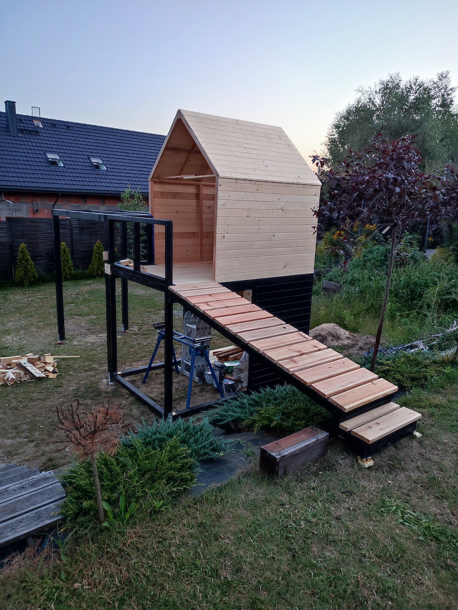Niedokończony drewniany domek dla dzieci na podwyższeniu z rampą, widoczne rusztowania i narzędzia, w tle dom z ciemną dachówką i zielona roślinność.