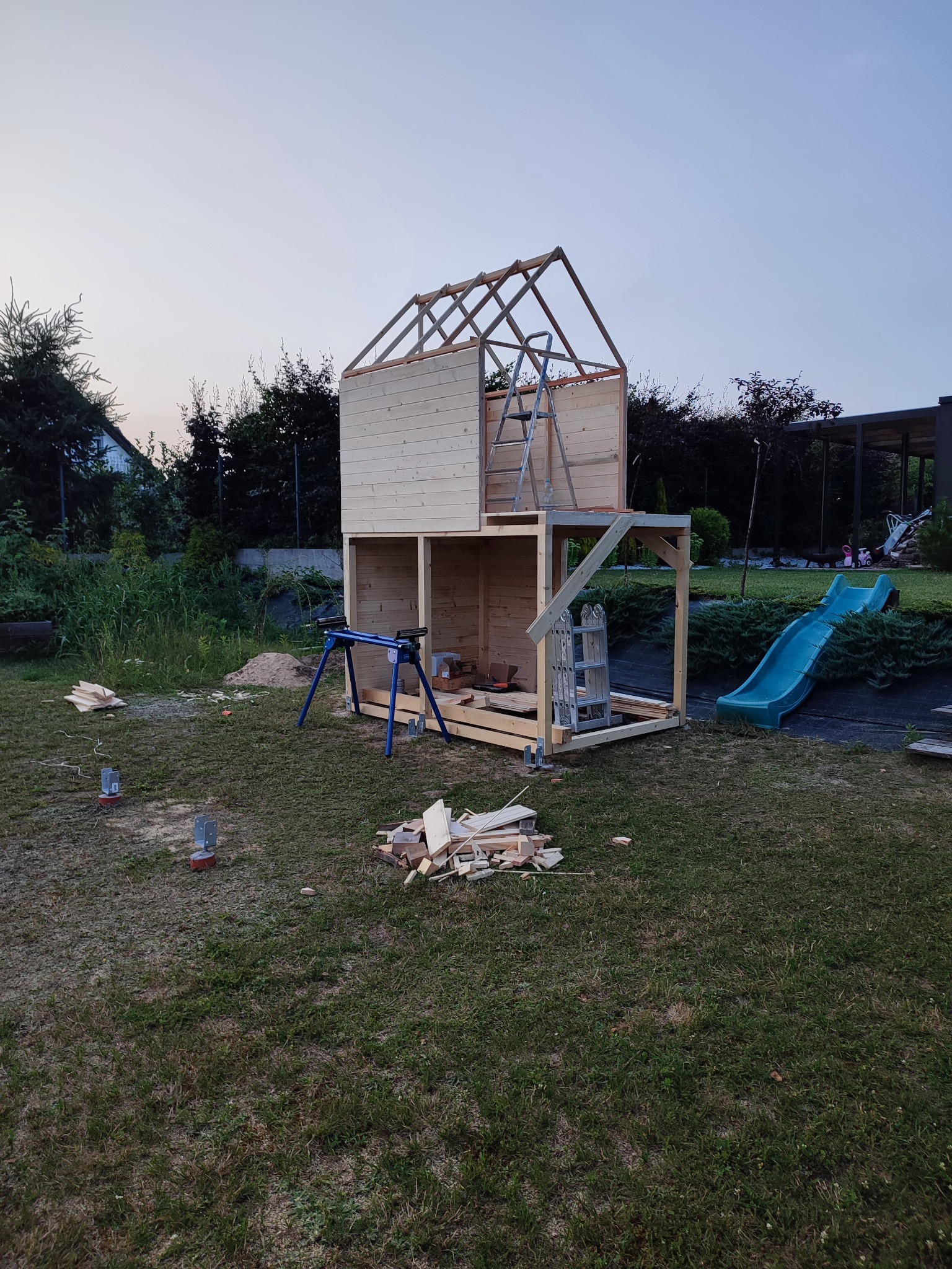 Drewniana konstrukcja domku dla dzieci w trakcie budowy na trawiastym podwórku, widoczna drabina i zjeżdżalnia, narzędzia i resztki drewna porozrzucane wokół.