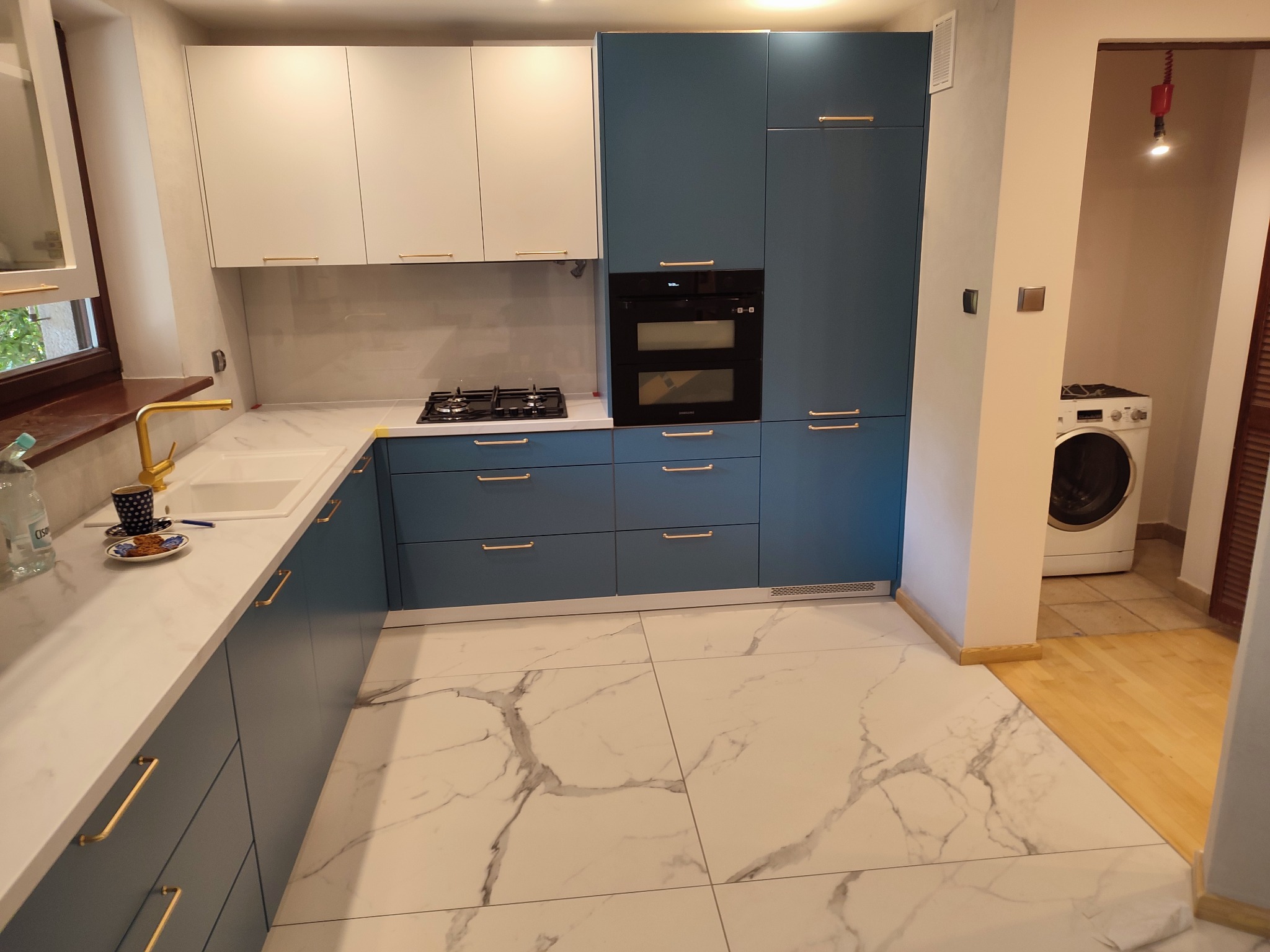 Nowoczesna kuchnia z białymi i niebieskimi szafkami, złotymi uchwytami, marmurową podłogą i złotą baterią; w tle widoczna pralka.