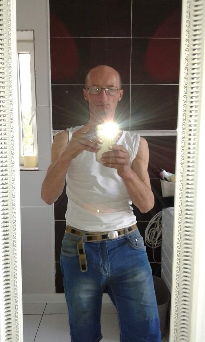 Mężczyzna w okularach robi selfie w lustrze łazienki, widoczne kafelki i grzejnik.