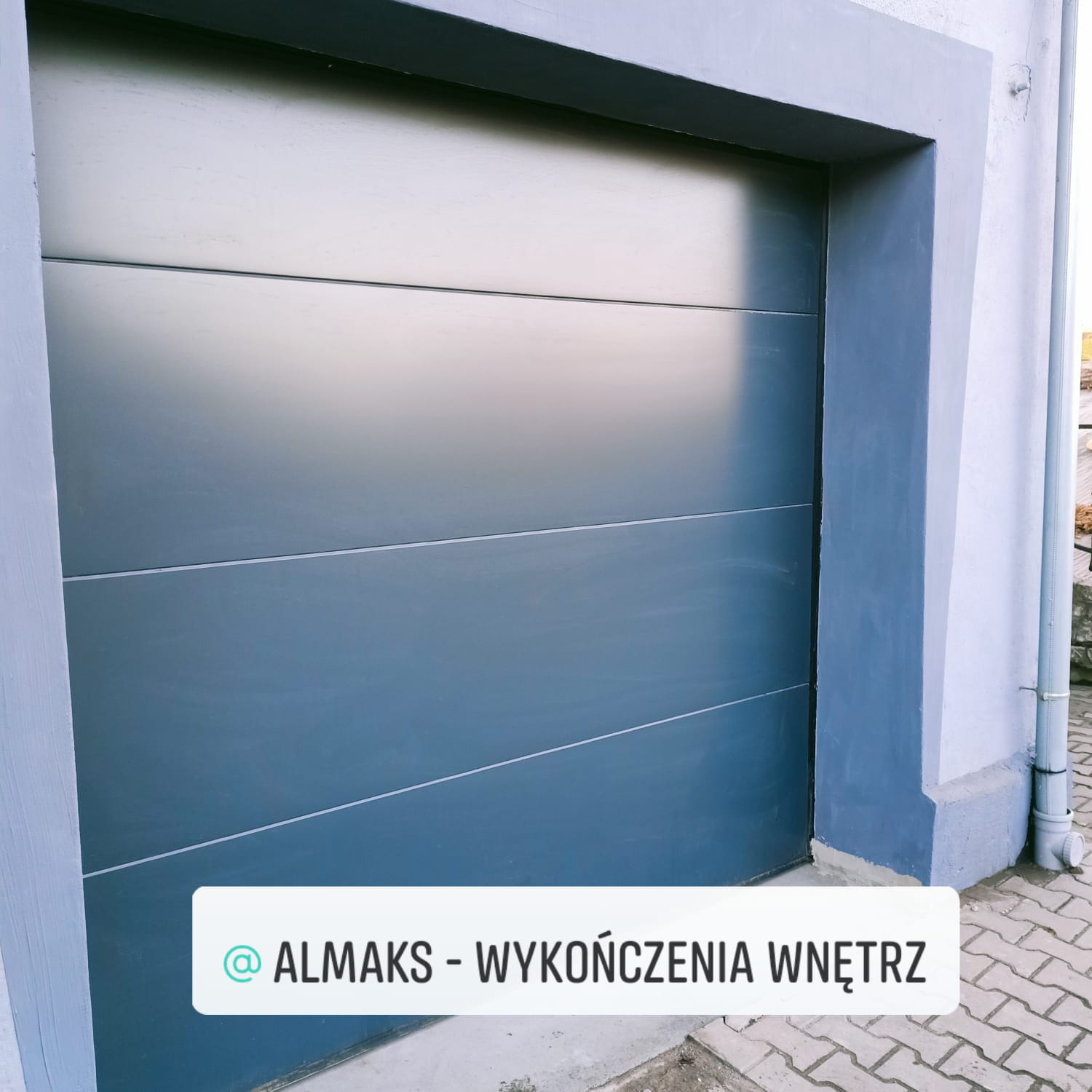 Szara segmentowa brama garażowa z czterema poziomymi panelami, zamontowana w szarej ramie, widoczny fragment chodnika z kostki brukowej i rynna spustowa.