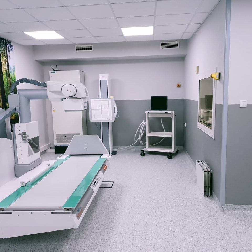 Nowoczesne, sterylne wnętrze gabinetu radiologicznego z aparaturą RTG, w tle tapeta z motywem lasu iglastego.