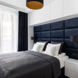 Nowoczesna sypialnia z tapicerowanym panelem ściennym w kolorze granatowym, białą zabudową meblową, zasłonami w odcieniach granatu i bieli oraz lampą wiszącą z elementami w kolorze złota.