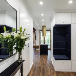 Nowoczesny przedpokój z białą zabudową meblową, wnęką z granatową tapicerką, lustrem i bukietem białych lilii w wazonie na czarnym stoliku konsolowym.