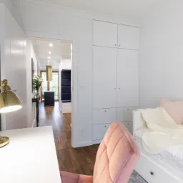 Biały pokój z łóżkiem, biurkiem z lampką w stylu retro i szafą wnękową, widok na korytarz z designerskim oświetleniem.