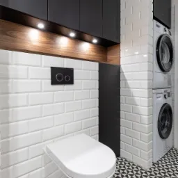 Nowoczesna łazienka z podwieszaną toaletą, białą cegiełkową glazurą, czarnymi szafkami i pralką z suszarką w kolumnie, oświetlenie punktowe nad drewnianym blatem i geometryczna posadzka.