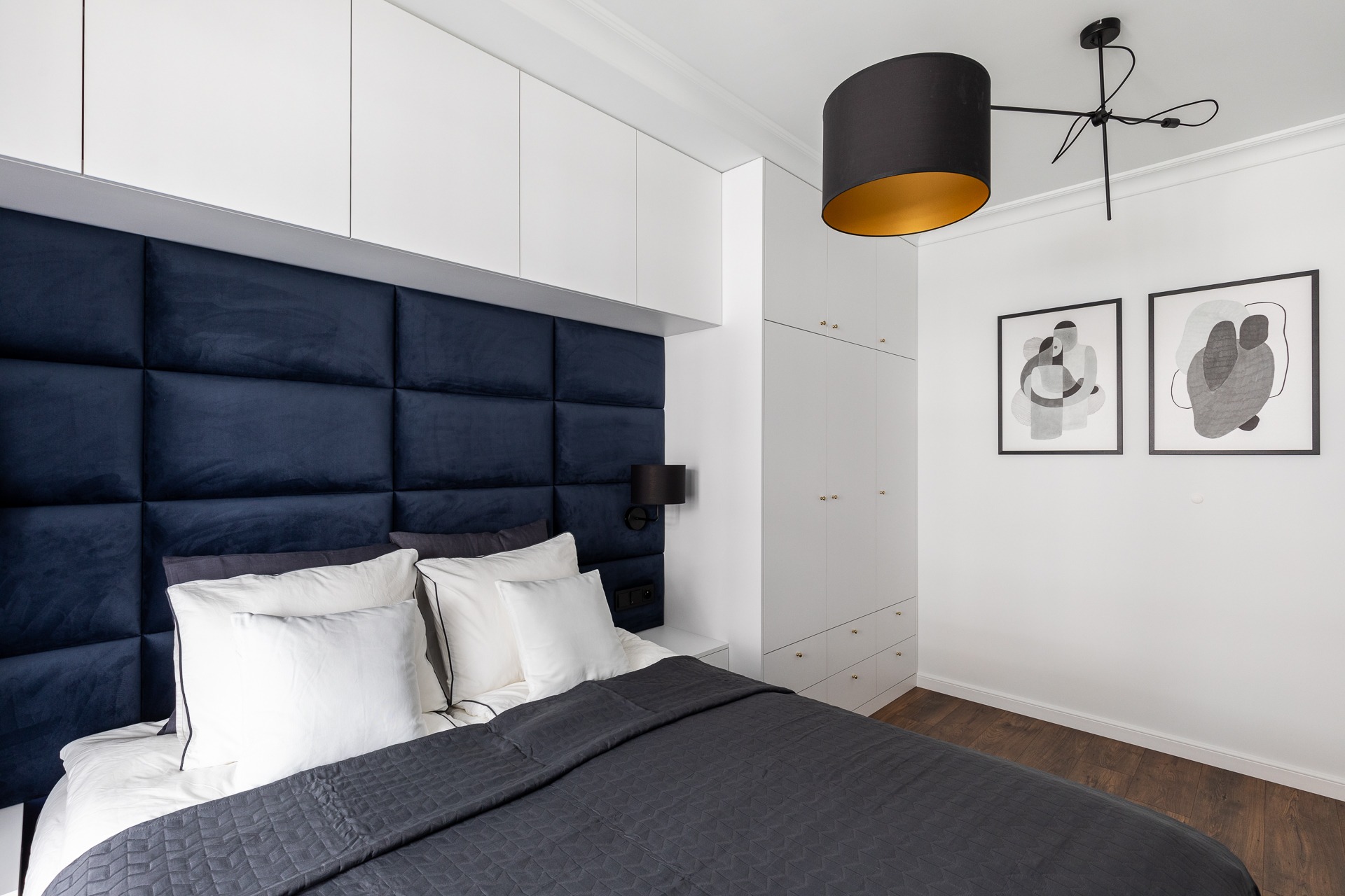 Nowoczesna sypialnia z tapicerowanym, granatowym wezgłowiem, białą zabudową szaf, minimalistyczną lampą i abstrakcyjnymi obrazami na ścianie.