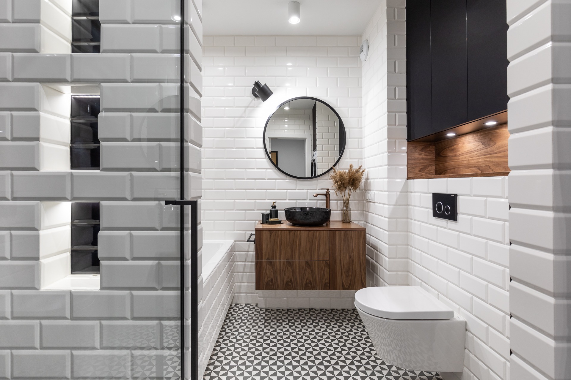 Nowoczesna łazienka z białą cegiełką na ścianach, drewnianą szafką pod umywalką z czarną misą, okrągłym lustrem i geometryczną podłogą w czarno-białe trójkąty.