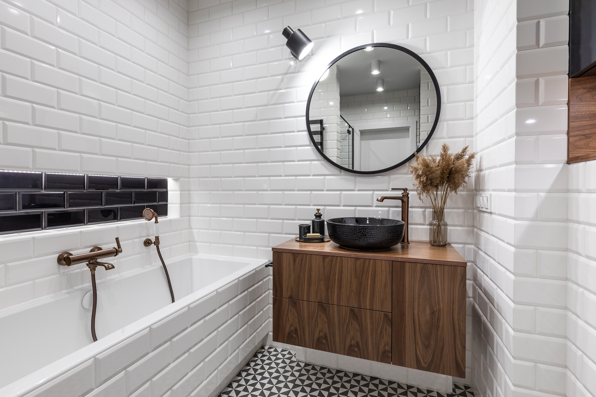 Nowoczesna łazienka z białą cegłą, czarnymi akcentami i drewnianą szafką pod umywalką z czarną, teksturowaną misą, bateria w kolorze miedzi, okrągłe lustro i geometryczna podłoga.