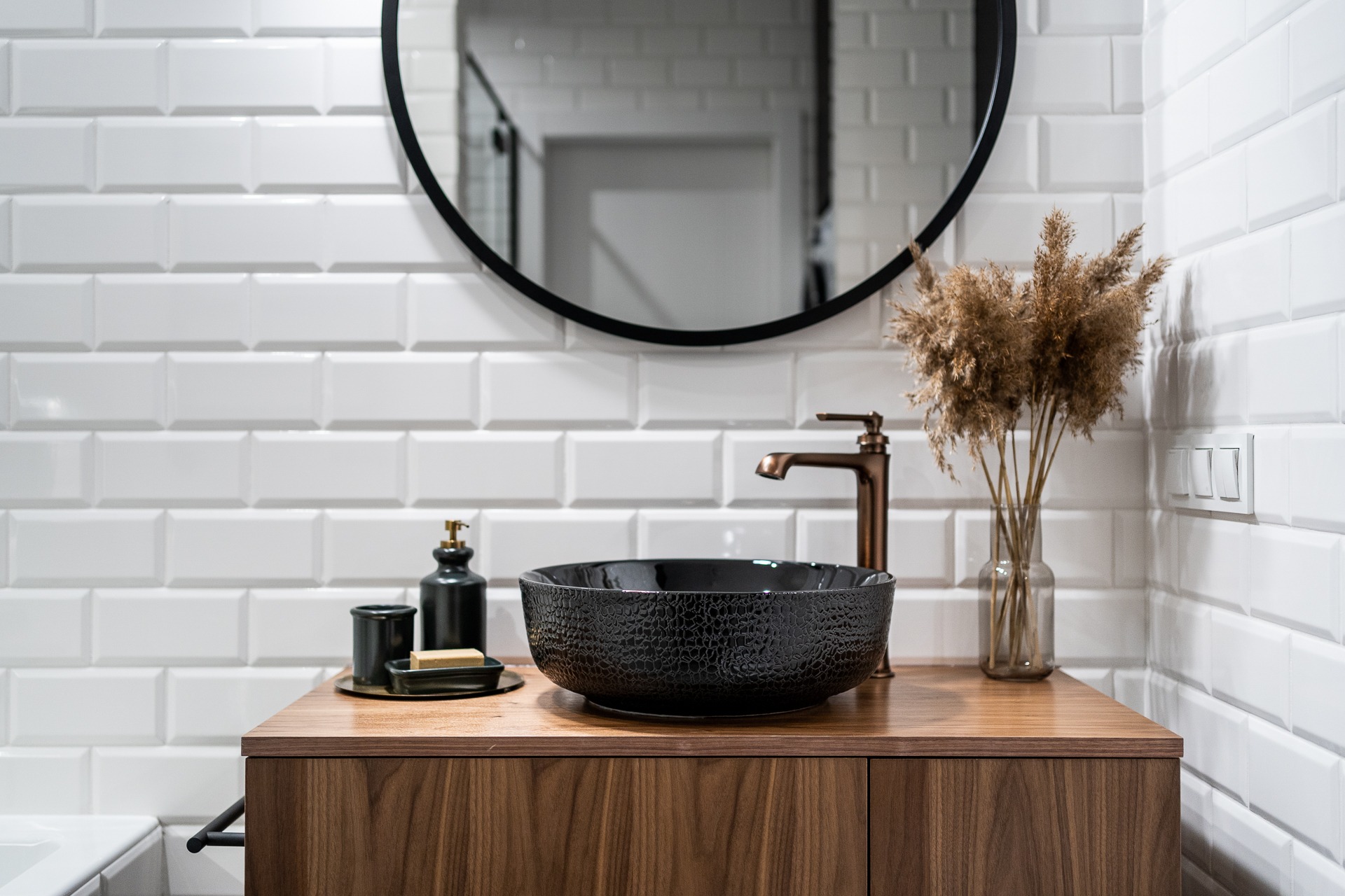 Nowoczesna łazienka z umywalką nablatową o fakturze skóry węża, baterią w kolorze miedzi, okrągłym lustrem i białymi płytkami typu metro. Dekoracyjny bukiet suszonych traw w szklanym wazonie.