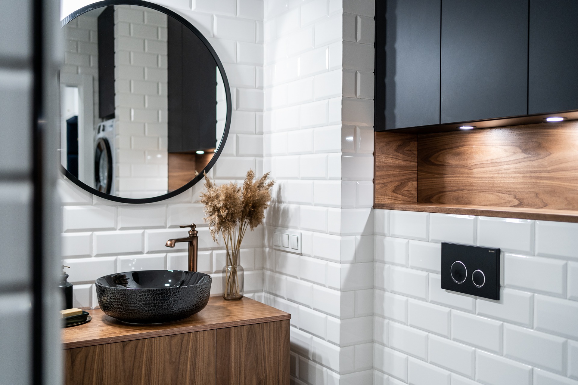 Nowoczesna łazienka z umywalką nablatową w kolorze czarnym o fakturze skóry, okrągłym lustrem w czarnej ramie, baterią w kolorze miedzi i białymi płytkami typu metro, uzupełniona drewnianą szafką...