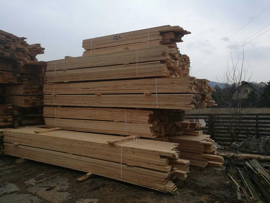 Stosy świeżo przetartego drewna budowlanego, pakiety związane sznurkiem, widoczne oznaczenia '124' na górnym pakiecie, w tle zabudowania wiejskie i pochmurne niebo.