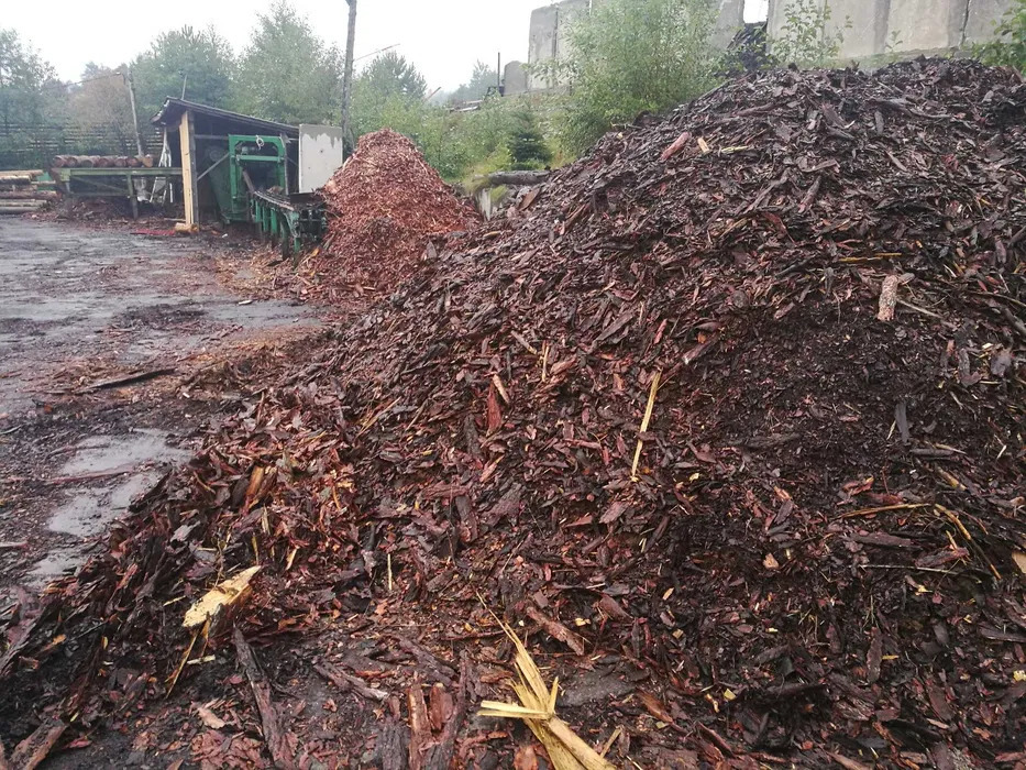 Duże pryzmy ciemnych trocin i zrębków drzewnych na placu tartaku, widoczna maszyna do rozdrabniania drewna w tle.