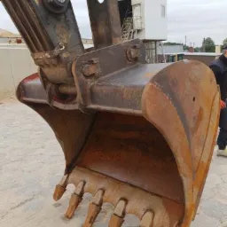 Bliskie ujęcie łyżki koparki z widocznymi zębami, z elementami konstrukcyjnymi i fragmentem białego budynku w tle.