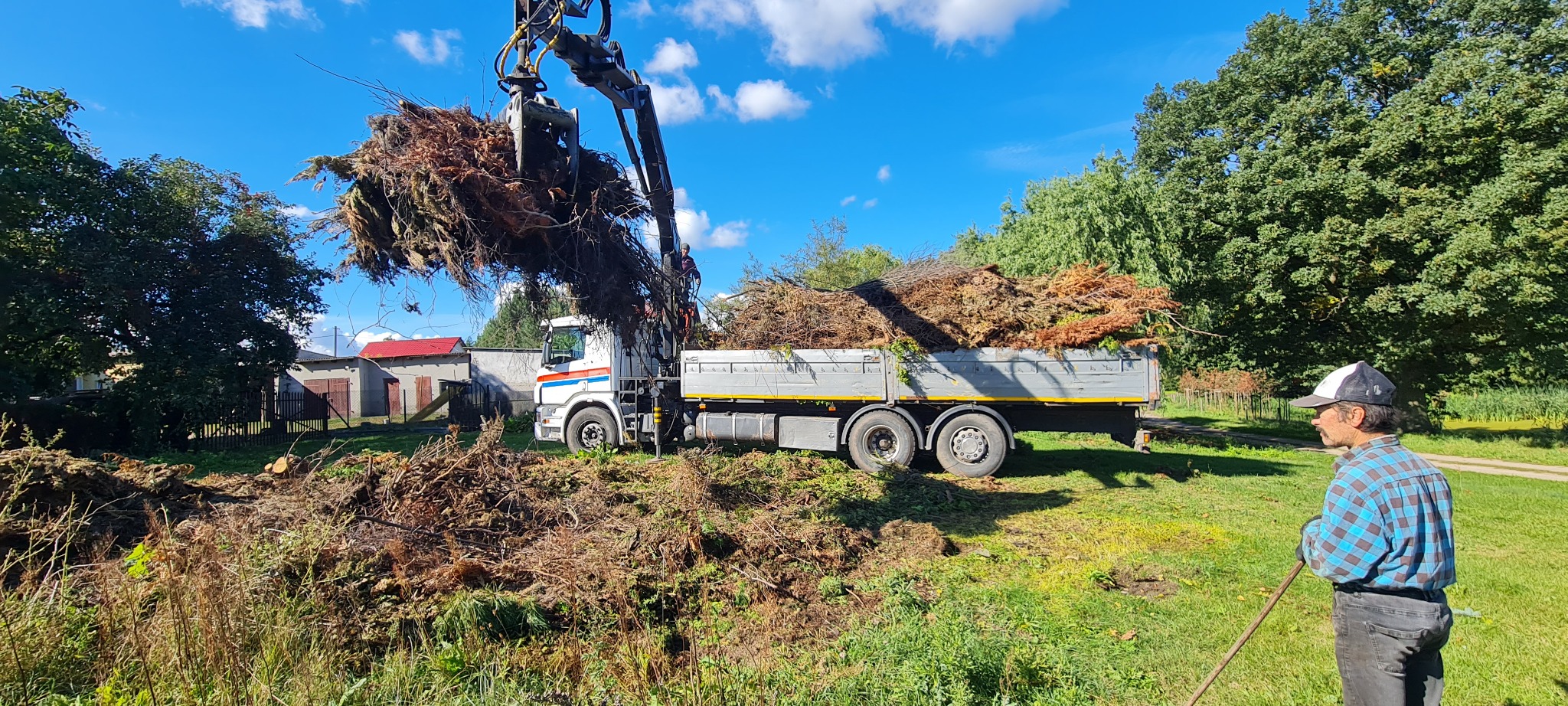 Ciężarówka z podnośnikiem załadowywana gałęziami i resztkami roślin po wycince na tle zieleni, pracownik w czapce z grabiami obserwuje proces.