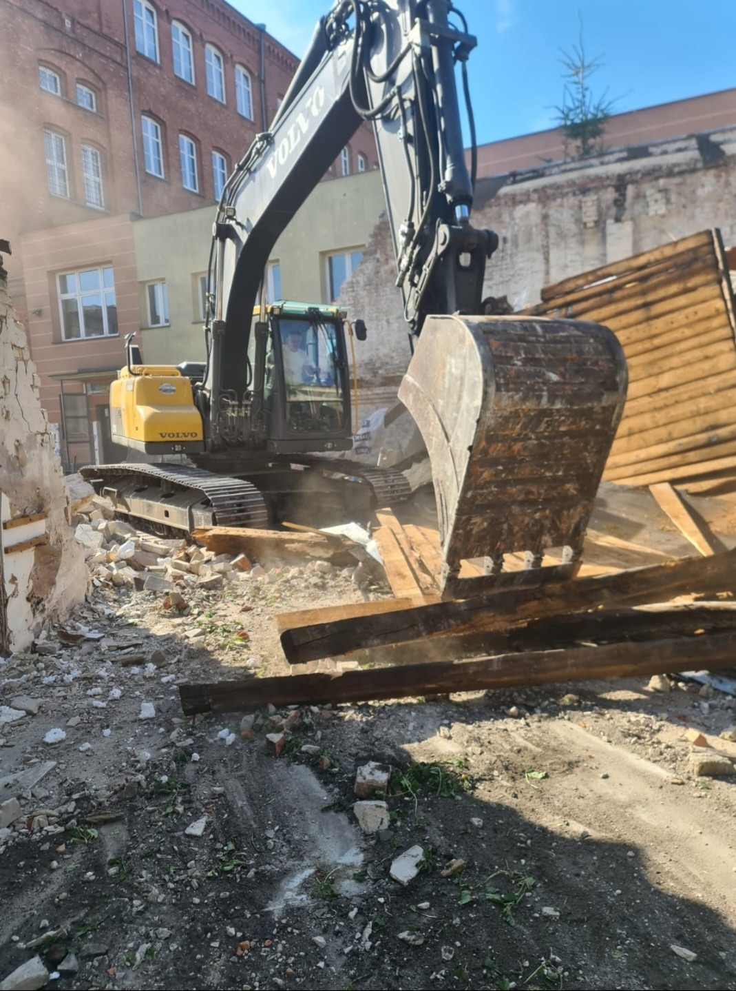 Żółta koparka Volvo z dużą łyżką kruszy resztki muru i drewniane elementy podczas rozbiórki budynku w miejskiej zabudowie, widoczne ceglane budynki w tle.