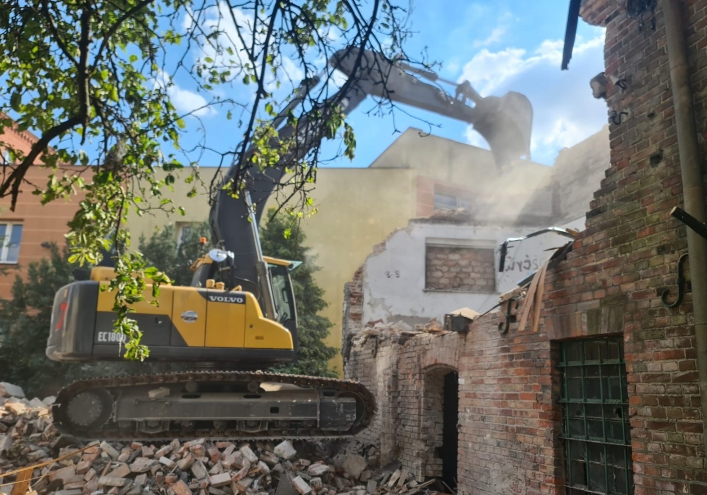 Żółta koparka Volvo EC180 na gruzowisku ceglanego budynku w trakcie rozbiórki, z widocznymi fragmentami zieleni i błękitnego nieba w tle.