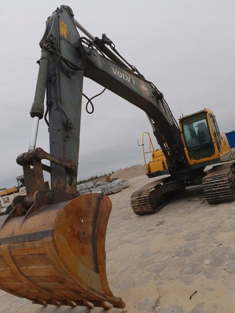 Żółto-szara koparka Volvo na placu budowy, widok z dołu na łyżkę i ramię maszyny, w tle materiały budowlane i pochmurne niebo.