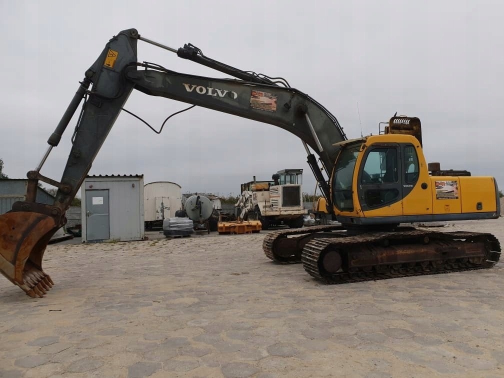 Żółto-szara koparka Volvo na placu budowy, z widocznymi śladami użytkowania na gąsienicach i łyżce, w tle barak i inne maszyny.