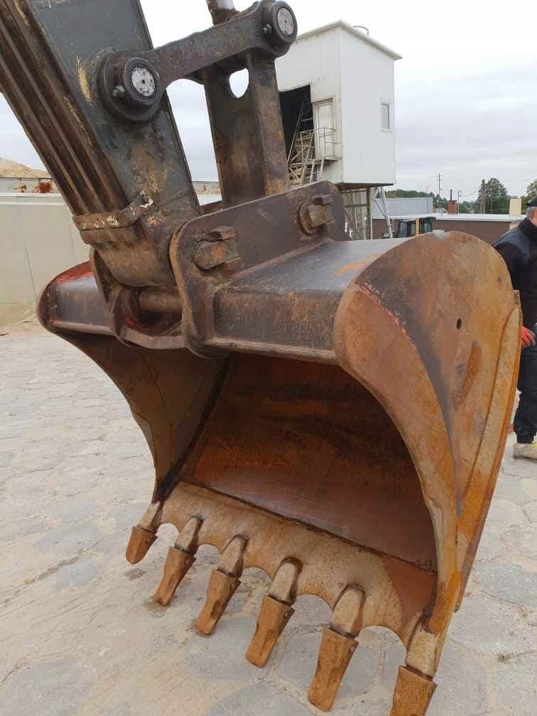 Bliskie ujęcie łyżki koparki z widocznymi zębami, z elementami konstrukcyjnymi i fragmentem białego budynku w tle.