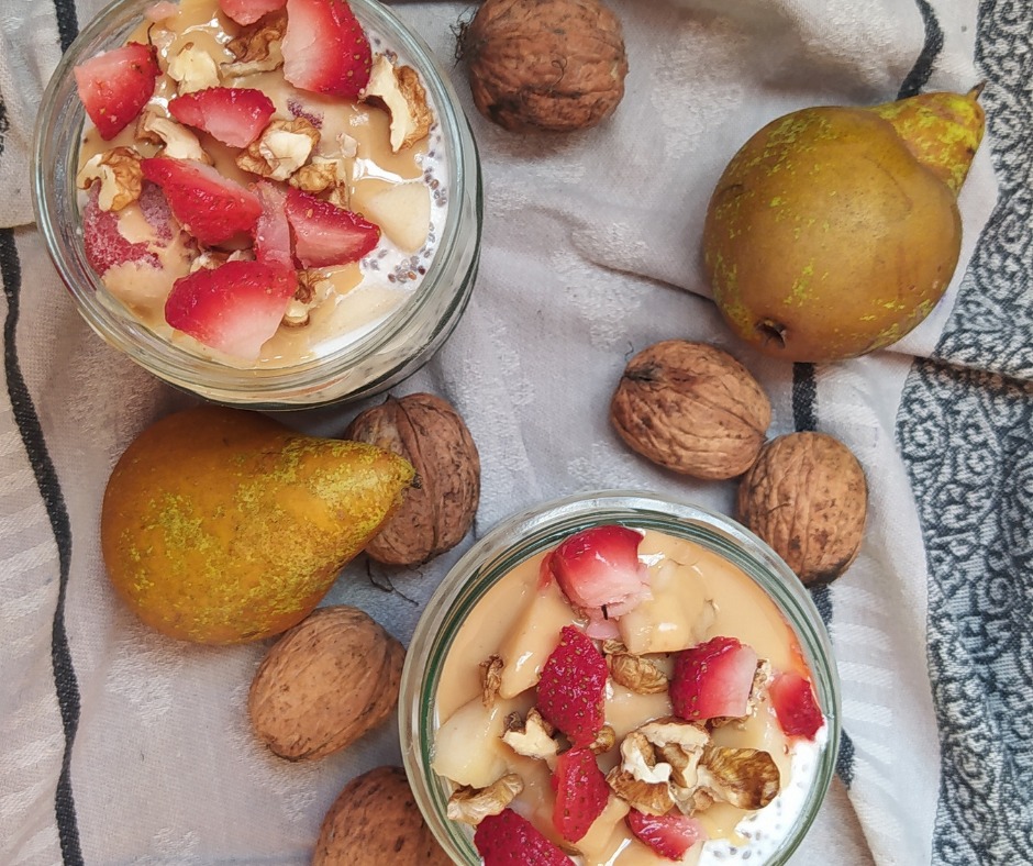 Dwa słoiki z deserem chia z kawałkami truskawek, gruszek i orzechów włoskich, otoczone dodatkowymi gruszkami i orzechami na wzorzystej szarej ściereczce.