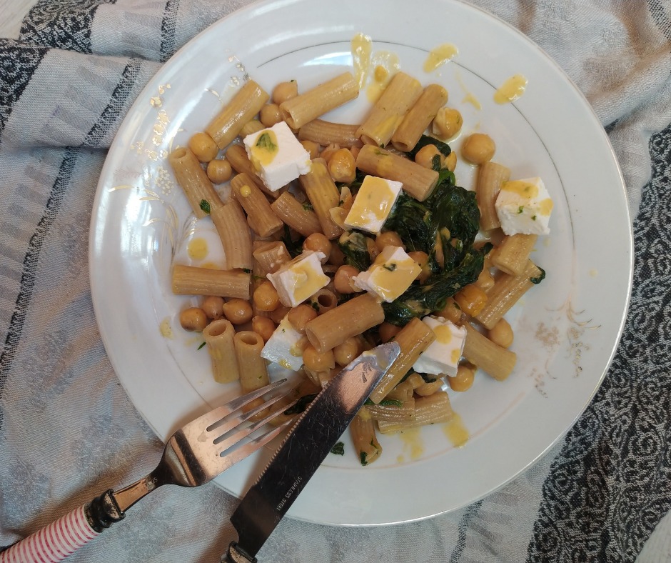 Makaron rigatoni z ciecierzycą, szpinakiem i kostkami sera feta na białym talerzu z widelcem i nożem, na wzorzystej ściereczce.