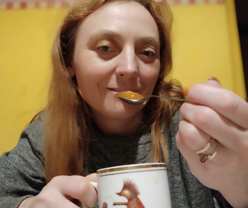 Uśmiechnięta kobieta z łyżką zupy w dłoni, zbliżenie na twarz i łyżkę z płynną pomarańczową zawartością, w tle żółta ściana i fragment kubka z rysunkiem.
