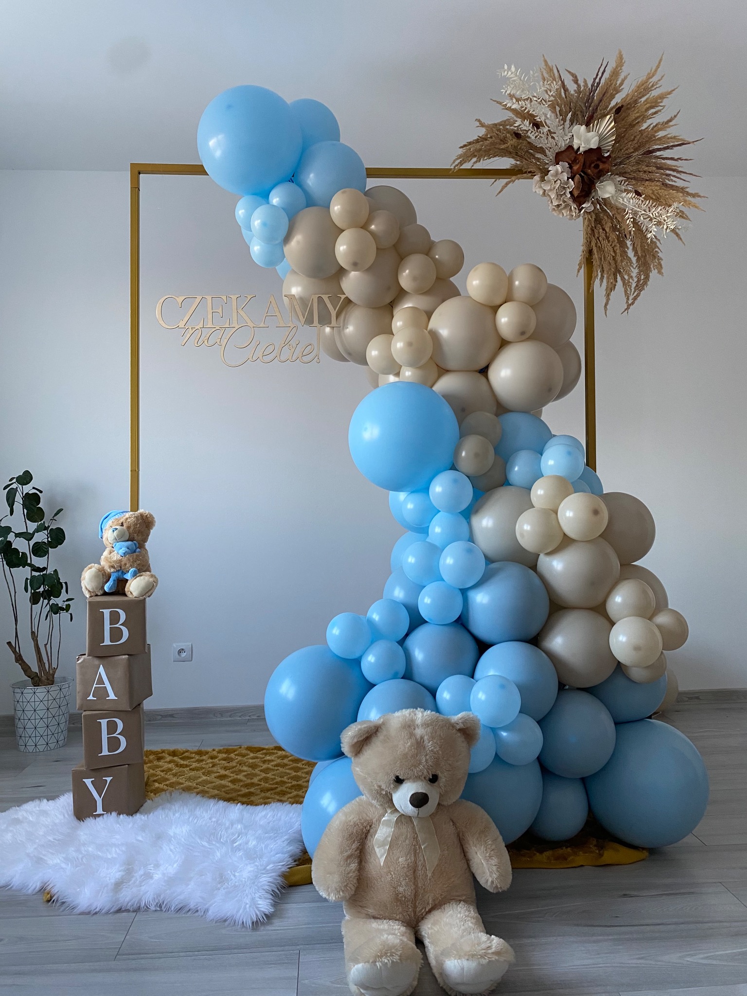 Dekoracja na baby shower: niebiesko-beżowa girlanda balonowa na złotej ramie, napis 'Czekamy na Ciebie', pluszowy miś, drewniane klocki z napisem BABY i pampasowa kompozycja kwiatowa.