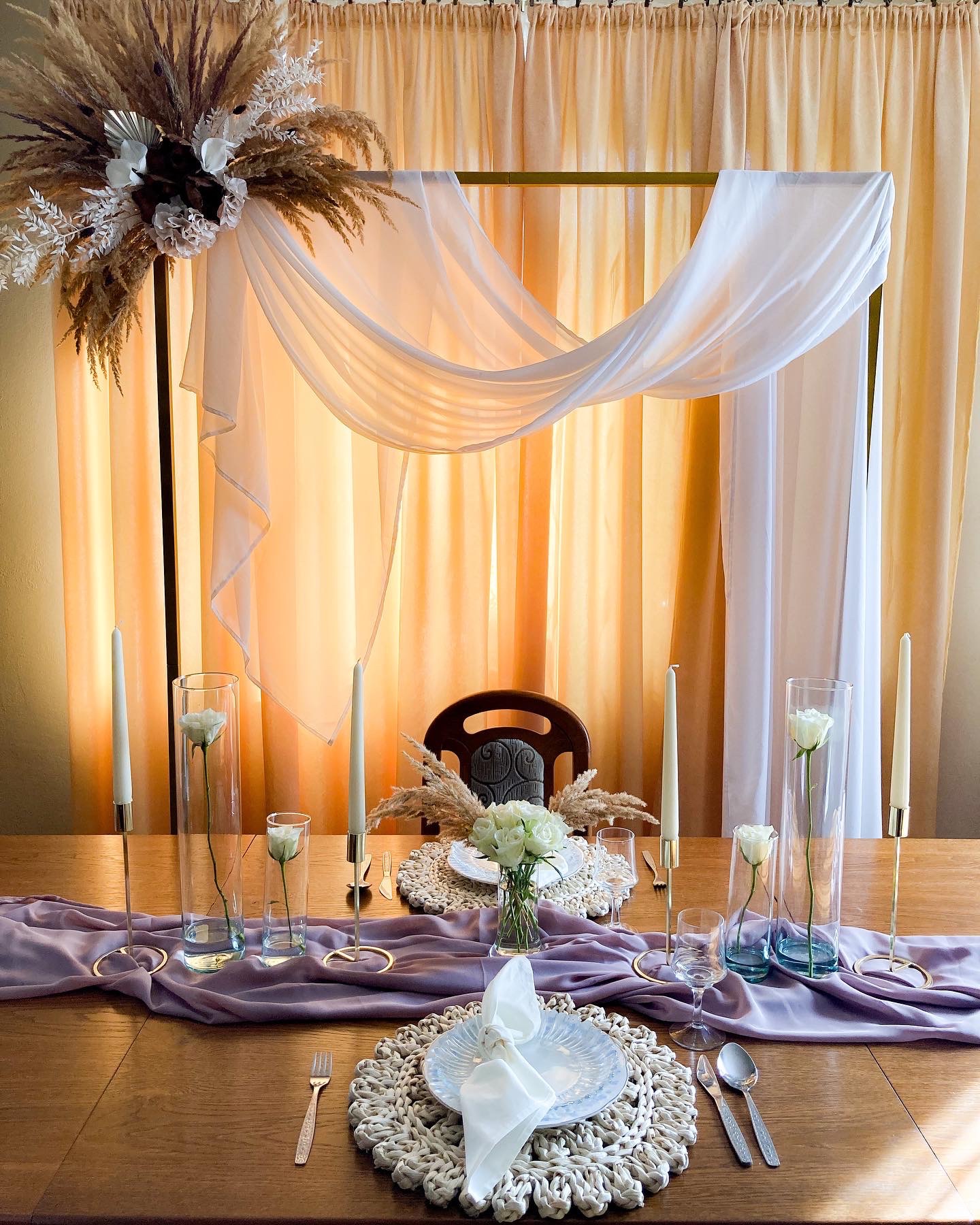 Uroczysta dekoracja stołu z kwiatami w wazonach, świecami w złotych świecznikach, beżowymi podkładkami i fioletowym bieżnikiem na drewnianym stole, tło stanowi złota konstrukcja z drapowanym białym...