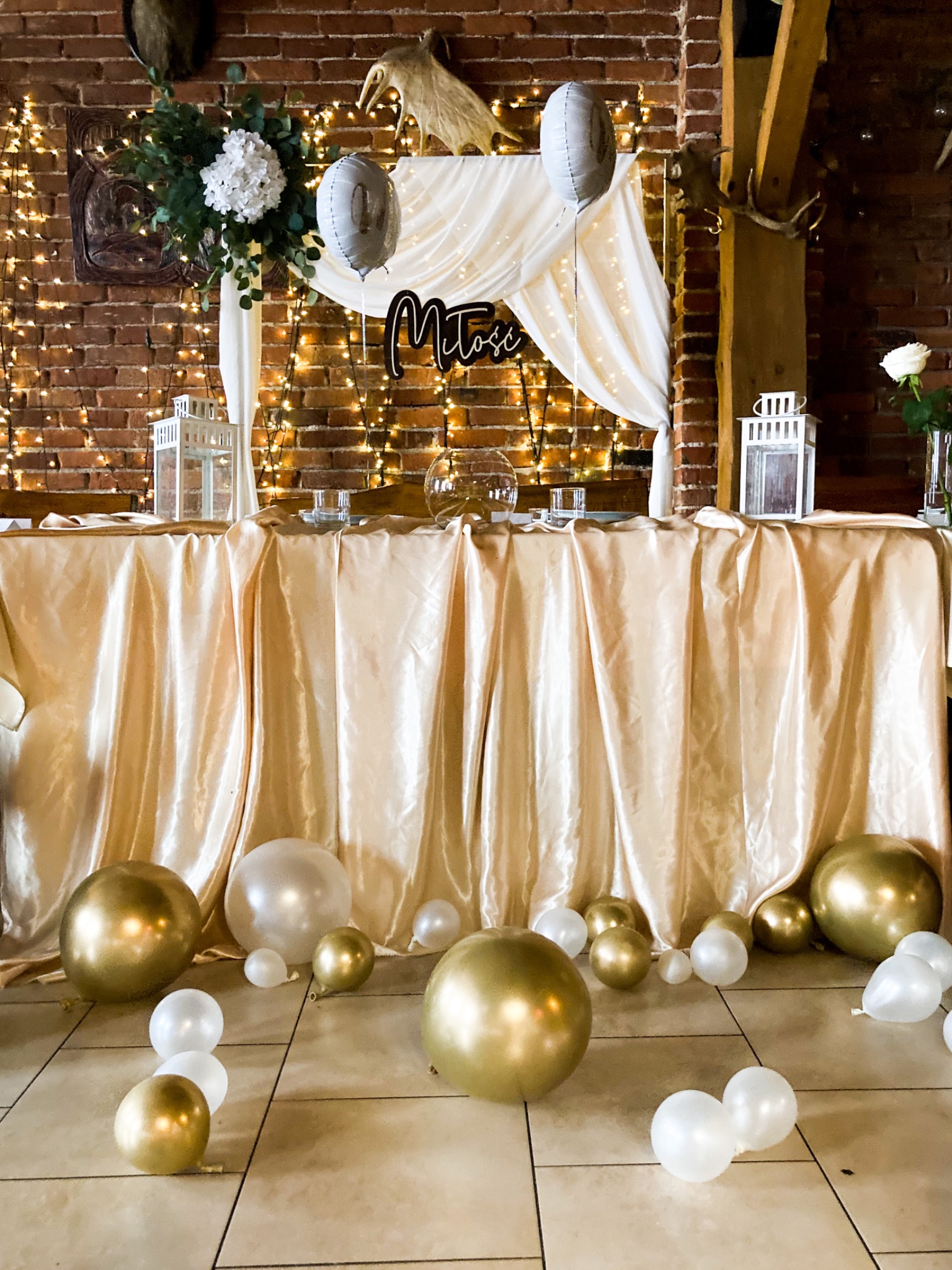 Dekoracja stołu na uroczystość z białymi i złotymi balonami, lampionami, kwiatami i napisem 'Miłości' na tle ceglanej ściany z lampkami.