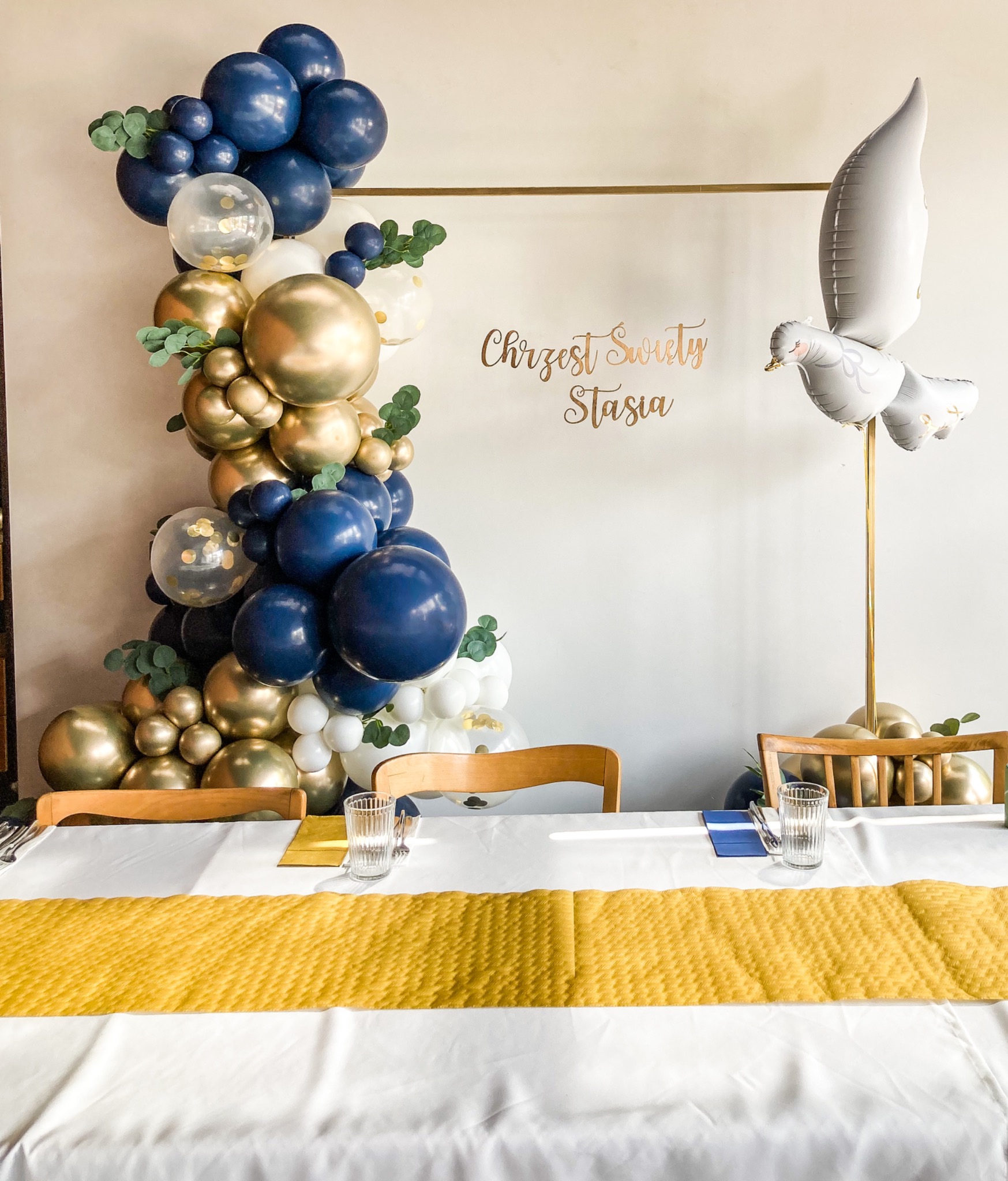 Dekoracja stołu na uroczystość Chrztu Świętego Stasi: girlanda z granatowych, złotych i białych balonów z zielonymi akcentami, balon w kształcie gołębia, biały obrus z żółtym bieżnikiem, nakrycia...