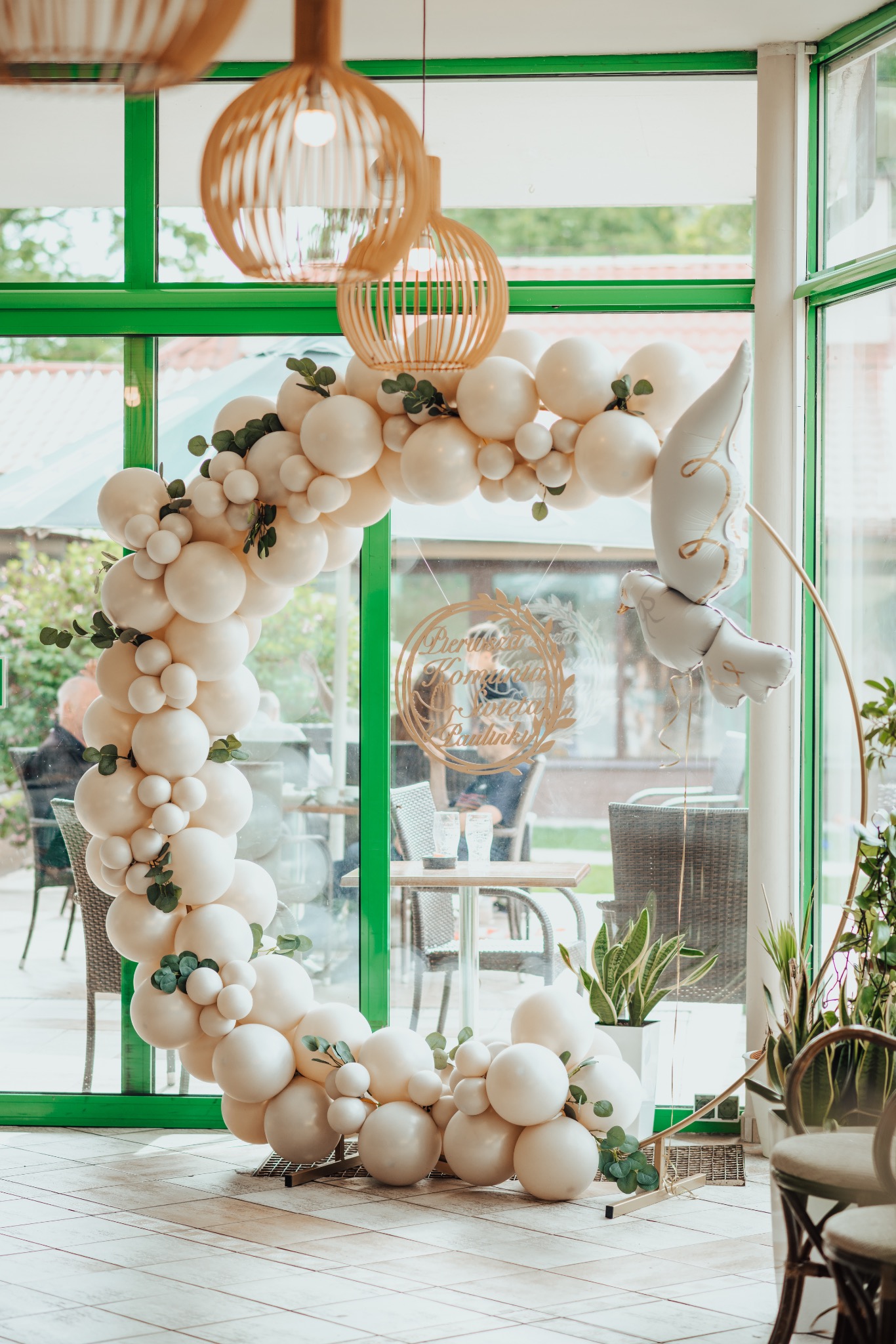 Dekoracja balonowa w kształcie półkola z zielonymi listkami i balonami w kształcie gołębi, ustawiona przy zielonych oknach lokalu we Wrocławiu, widoczna dekoracja z napisem 'Pierwsza Komunia Święta...
