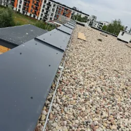 Widok z góry na dach pokryty drobnymi kamieniami z zamontowaną instalacją odgromową z szarą listwą i metalowym przewodem, na tle budynków mieszkalnych.