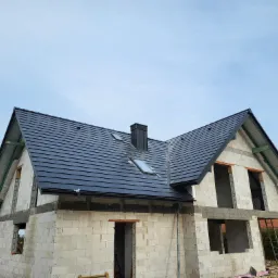 Surowy budynek jednorodzinny w trakcie budowy, z czarną dachówką i kominem, widoczne pustaki i niezabudowane otwory okienne, na tle błękitnego nieba.