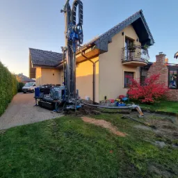 Wiertnica na posesji podczas wykonywania odwiertu, widoczne węże odprowadzające urobek, w tle dom jednorodzinny i zaparkowany biały samochód dostawczy.