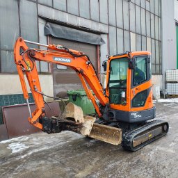 Firma Handlowo-Produkcyjno-Uslugowa 'Kocjan' - Pomarańczowa mini koparka Doosan DX35Z na tle hali przemysłowej z widocznymi śladami błota i śniegu na ziemi.