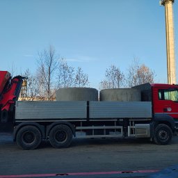 Firma Handlowo-Produkcyjno-Uslugowa 'Kocjan' - Czerwony samochód ciężarowy MAN z dźwigiem i dwoma betonowymi kręgami w skrzyni ładunkowej, na tle drzew i wysokiego komina.